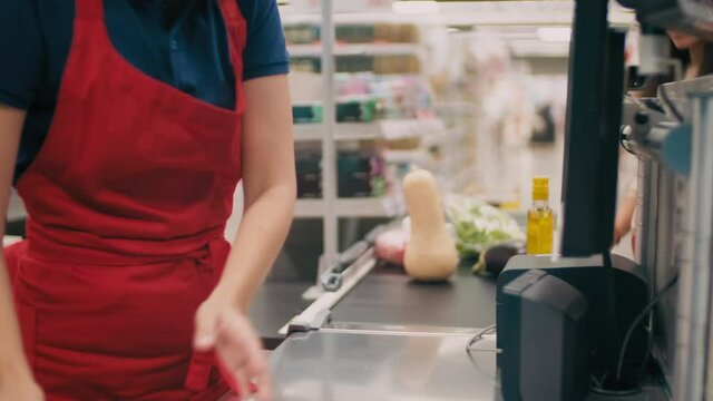 Midsection footage of unrecognizable woman checking out at big supermarket and cashier in red uniform weighing vegetables