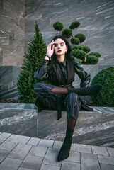 Young girl model in fashionable clothes on background of black stone wall and green plants,...