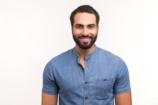 Positive handsome bearded man in blue shirt looking at camera with toothy smile, happy successful business man. Indoor studio shot isolated on white background