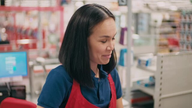 Medium close-up of pretty mixed-race cashier talking to customers smiling while scanning items at counter in supermarket