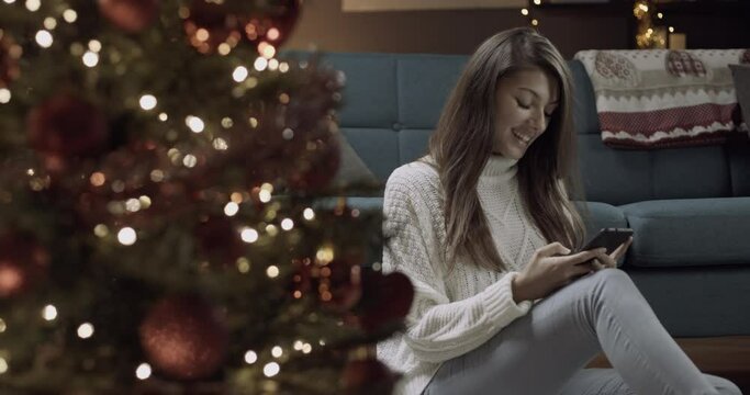 Young woman typing on a smartphone sitting near christmas tree and gifts.