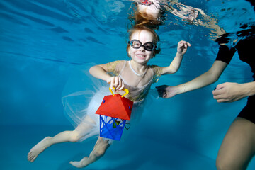 Obraz na płótnie Canvas Photo of a laughing little girl under the water in the pool. She's holding a flashlight. Baby learns to dive. Swimming lessons with a child. Healthy lifestyle. Horizontal orientation