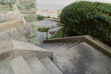 Concrete stairs descending around an outside corner of a building made from crumbling green limestone, black handrails on steep descent, horizontal aspect