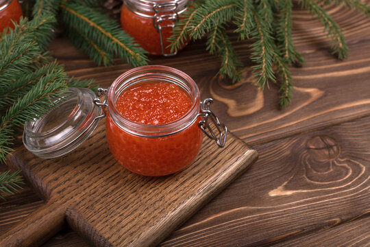Red caviar in a glass jar on a brown wooden background with a Christmas tree.