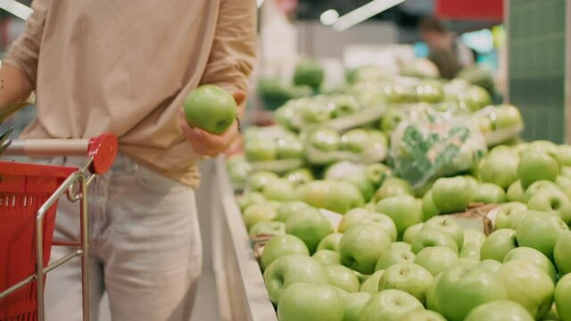 Midsection footage of woman with product cart doing shopping picking fresh green apples in big grocery store