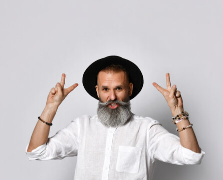 Portrait Of A Cheerful Middle-aged Man In A Fashionable Black Hat Standing On A Gray Background And Showing A Peace Sign. Street Style. Concept Of Stylish Older People. Place For Text.