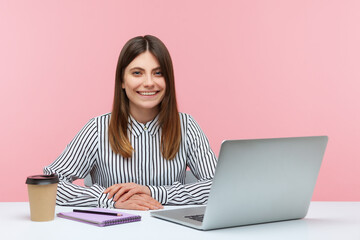 Happy cheerful office woman in striped shirt sitting workplace looking at camera with toothy smile, drinking coffee and making notes working on laptop. Indoor studio shot isolated on pink background