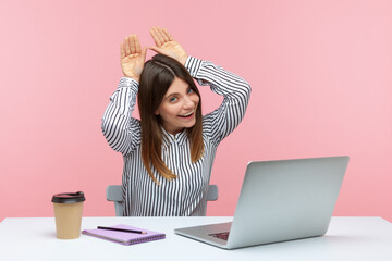 Positive woman office worker smiling showing bunny ears with hand on head, resting, having fun at workplace in home office, feeling carefree and happy. Indoor studio shot isolated on pink background