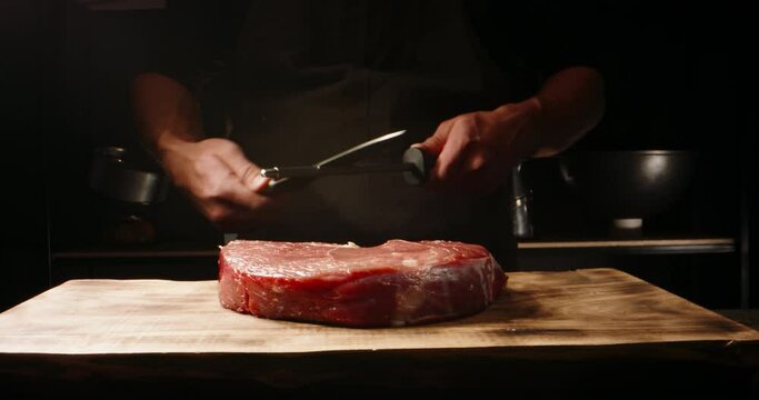 Chef sharpening his knife in front of raw piece of steak. Cooker preparing his tools for cutting meat for grilling on professional kitchen table 4k footage
