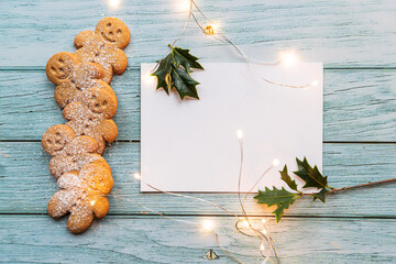 gingerbread cookies, gingerman, arranged on a blue table with holly, traditional Christmas plant, Christmas lights, and a blank white card with copyspace, lets you create a tasty Christmas card - 397684957
