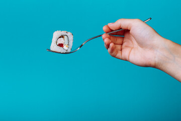 Hand holding fresh sushi roll with a fork, isolated on blue background. How to eat sushi without chopsticks
