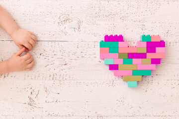 Top view of a colorful heart made of plastic cubes and a kid's hand on a white wooden background. A creative idea from bright colored cubes of a girl's color of the constructor. Early learning