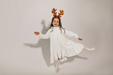 Full-lenght studio shot of little girl wearing white dress and Christmas accessories in the head dancing and having fun over isolated background