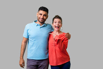 Obraz premium That's him! Portrait of happy young couple in casual wear standing and hugging as friends, woman pointing finger at man, looking shocked at camera. isolated on gray background, indoor studio shot