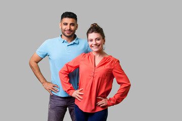 Portrait of confident attractive young couple in casual clothing standing together, holding hands on hips, smiling at camera, looking proud and happy. isolated on gray background, indoor studio shot