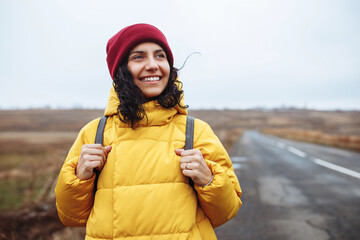 Portrait of a female tourist with a backpack wearing yellow jacket and red hat stands on the road. Young woman travels during winter or late autumn season. Hitch-hiking, trip and travelling concept.