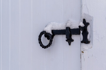 A white wooden door closed with the use of a vintage black wrought iron latch. The mechanism has a round handle. There's fresh white snow on the top of the old handle.  The shed building is white.