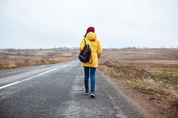 Backview of a female tourist with a backpack wearing yellow jacket and red hat walks on the road. Young woman travels during winter or late autumn season. Hitch-hiking, trip and travelling concept.