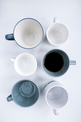 several different gray dark blue white glasses for tea or coffee stand on a white table. Only one cup is filled with coffee. Top down flat lay