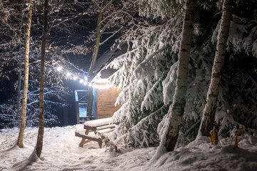 The charming view creates an incredible winter Christmas mood. A large fluffy spruce tree and a bench with a table near wooden lodge, everything covered with fluffy snow and lit by light from garlands