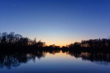 Clear blue sky after sunset at lake with reflecting silhouettes of trees