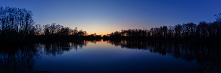 Clear blue sky after sunset at lake with reflecting silhouettes of trees panorama