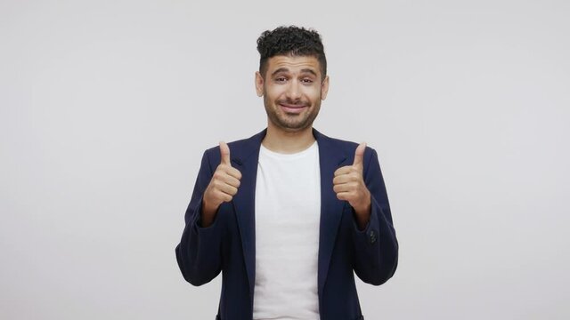 Positive satisfied bearded man in elegant suit showing thumbs up, looking at camera with happy excited expression, approving work, likes posts. Indoor studio shot isolated on gray background
