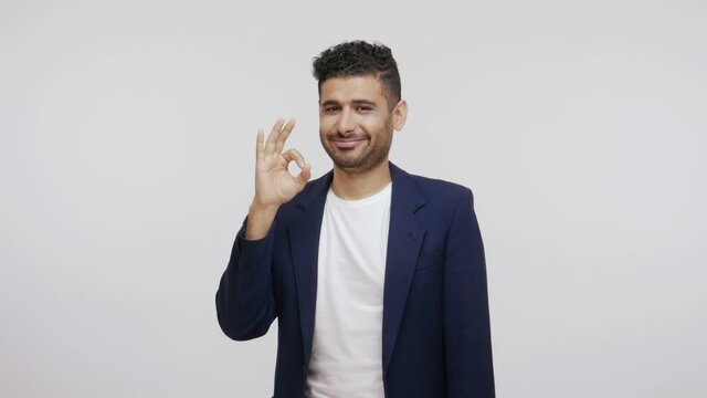 Happy extremely satisfied man with beard in suit showing ok gesture, approving work, proud and enjoying with good results, success. Indoor studio shot isolated on gray background