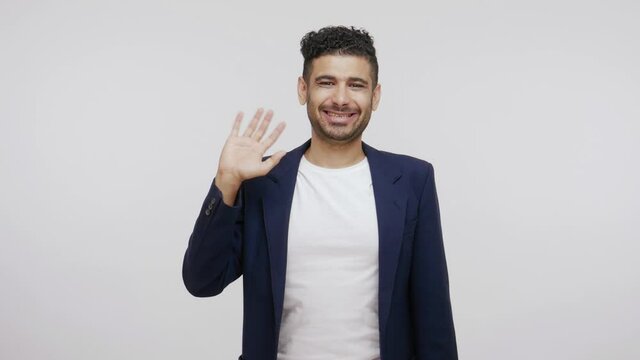 Extremely happy friendly man in suit waving hand looking at camera with smile, glad to see you, saying hi or good bye. Indoor studio shot isolated on gray background