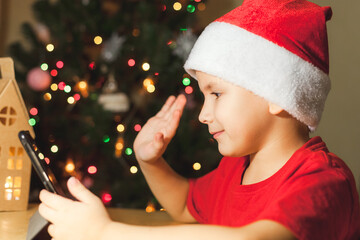 Cute little boy in red Christmas hat has video call using tablet. Decorated Christmas tree on background