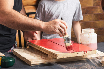 Father and son together make a wooden birdhouse in the workshop. Cheerful father with a little boy paint a wooden board with a brush in red  on a table in the workshop