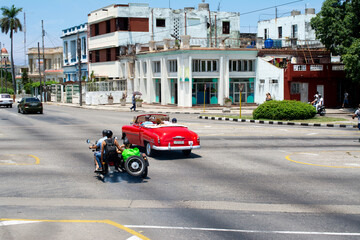En una avenida de La Habana hay varias personas que viajan en una motocicleta fabricada en Rusia con su sidecar y un coche clásico rojo.