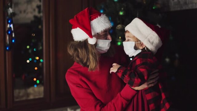 coronavirus, cavid-19 in the new year. a little daughter and her mother in a red Santa Hat and a medical mask are sitting at home under the Christmas tree and hugging.