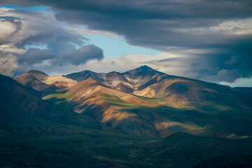 Fototapeta premium Awesome highland landscape with great mountains and blue clearance in cloudy sky in overcast weather. Blue skylight in cloudy sky above huge multicolor mountains in sunlight. Colorful mountain scenery