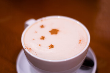 close-up of a white cup of coffee with froth decorated with cinnamon stars on a saucer with a spoon standing on a wooden table
