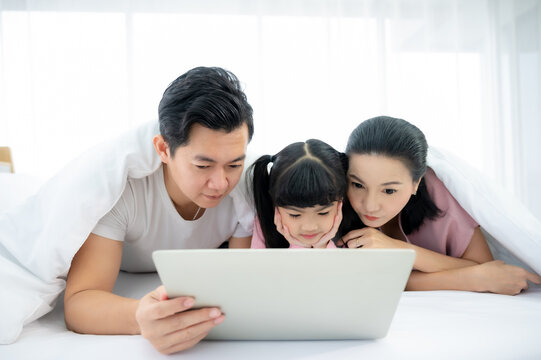 Happy asian family people leisure in bedroom together. Father and mother with daughter relaxing and using laptop on bed