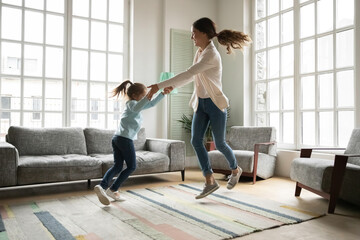 Full length overjoyed little kid girl holding mommy's hands, jumping twisting dancing to disco music together in modern living room, happy different generations family involved in funny activity.
