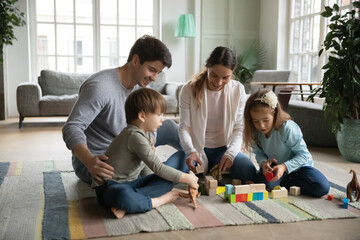 Affectionate caring young parents sitting on floor carpet with little joyful children siblings,...