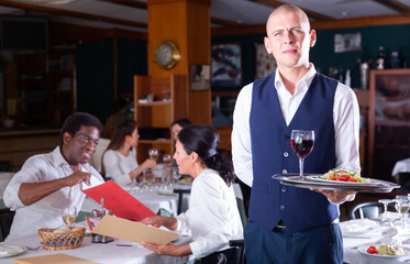 Fototapeta na wymiar Portrait of friendly headwaiter with serving tray warmly welcoming in cozy modern restaurant