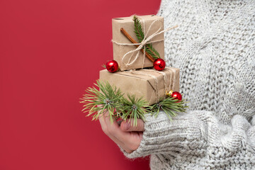 An unrecognizable girl in a sweater holds Christmas gifts on a red background. Handmade gift boxes in the hands of a woman. New year background with beautiful gifts