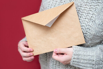 Brown envelope made of Kraft paper in women's hands on a red background, close-up. A letter to Santa, wish list concept. The girl is holding a letter. New year background