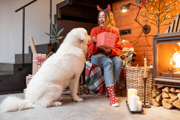 Young woman with her cute white dog unpacking gift boxes by a fireplace during a happy New Year holidays at home