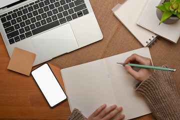 Female hand writing on blank notebook on worktable with laptop and smartphone