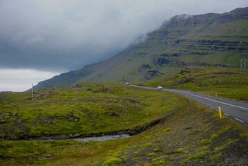 cars drive on paved road near green mountains, heavy low clouds in the sky, nature of Iceland