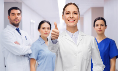 Fototapeta na wymiar medicine, profession and healthcare concept - happy smiling female doctor in white coat showing thumbs up over group of colleagues at hospital on background