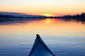 Bow of blue kayak against the background of a dawn, a quiet winter river, a dark blue water surface and a blue sky. Symbol of movement towards adventure. The kayak floats towards the sun.