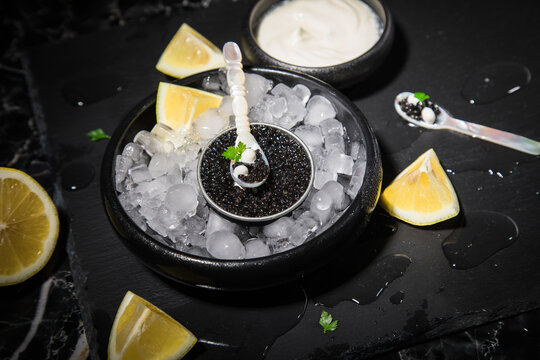 Schwarzer Störkaviar auf Eis mit Perlmutt Löffel, Schmand, Zitrone auf Schieferplatte und dunkel Marmor Hintergrund