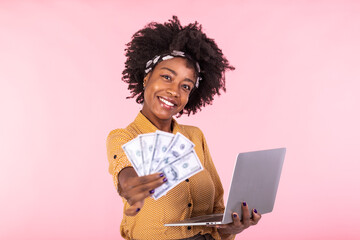 Image of cheerful African American woman standing isolated over pink background using laptop computer and holding money banknotes . Portrait of a smiling girl holding laptop computer