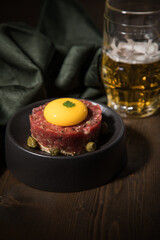Gourmet Rindertatar Beef Tatar roh aus Filet fettarm, Kapern, Eigelb, Zwiebeln auf Toast Brot und schwarz Teller mit Bier, grün Serviette und Holz Hintergrund dunkel
