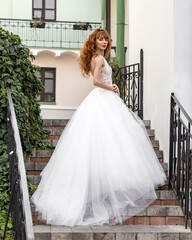 Mid adult bride in a voluminous dress with red curly hair stands on the steps of the stairs against the background of the old city. Exterior fashion shot of fashion model in wedding dress